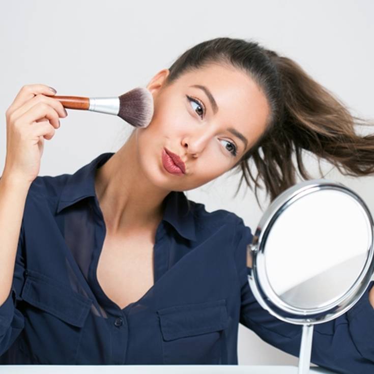 Imagem mostra uma mulher em frente ao espelho, aplicando blush em seu rosto.